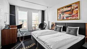 A modern hotel room with a large bed, a desk, a chair, and a view of the city through large windows. - Best Western Plus Hotel Eyde (Herning)