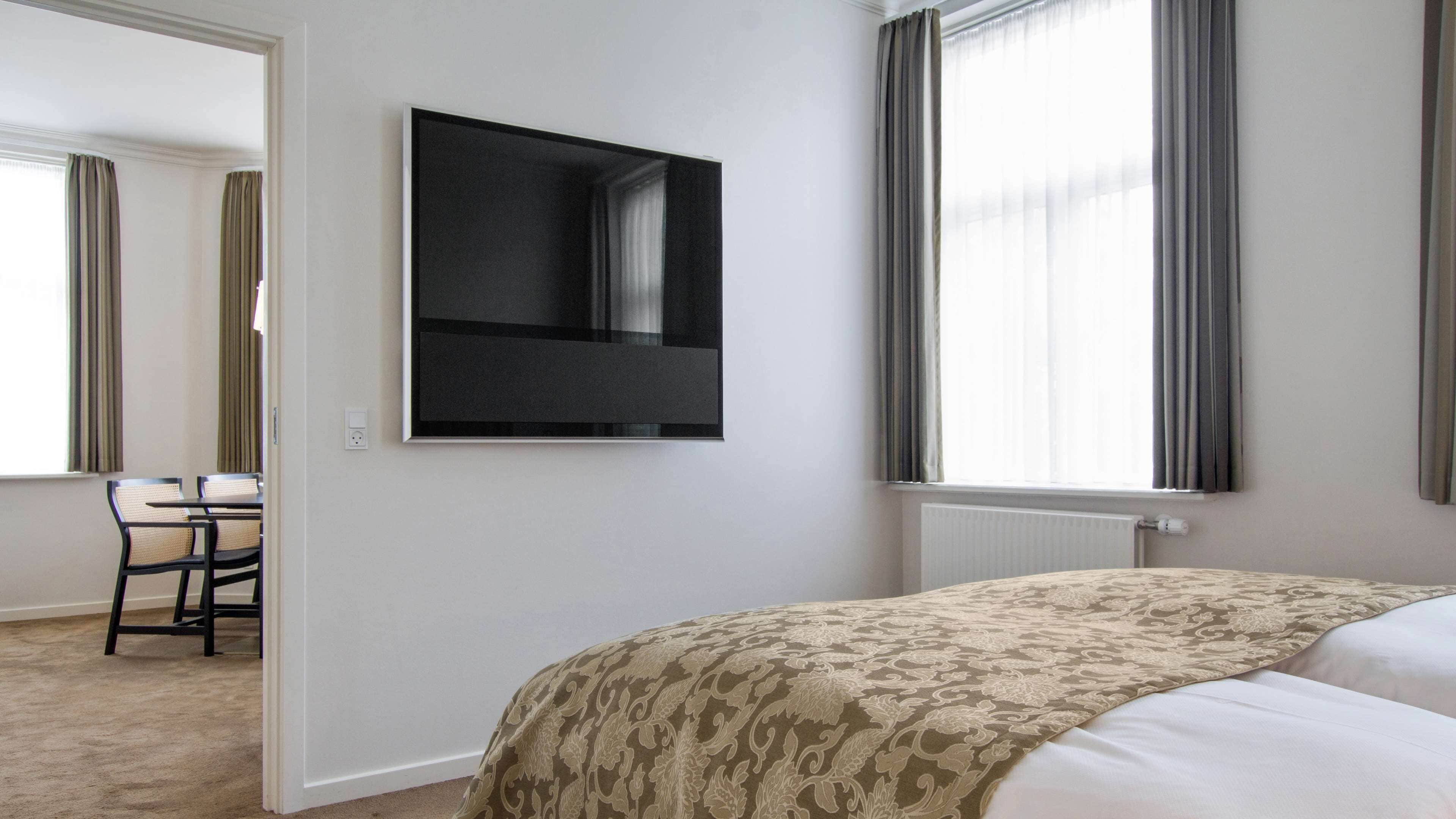 A hotel room with a bed, a TV mounted on the wall, a desk with chairs, and large windows with curtains.