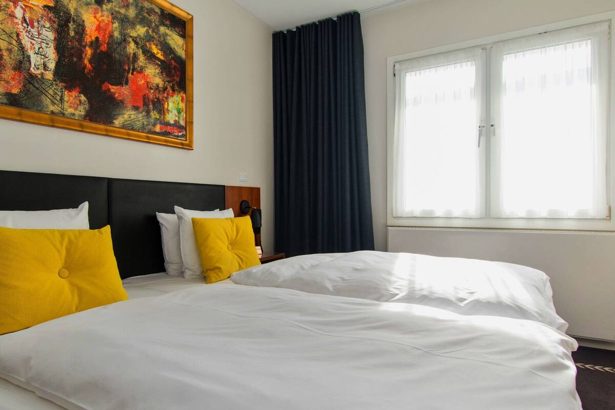 Habitación de hotel con dos camas, cada una con ropa de cama blanca y almohadas amarillas, un cuadro abstracto grande en la pared y una ventana con cortinas oscuras.