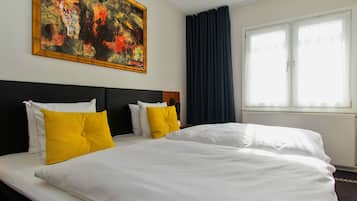 Habitación de hotel con dos camas, cada una con ropa de cama blanca y almohadas amarillas, un cuadro abstracto grande en la pared y una ventana con cortinas oscuras.