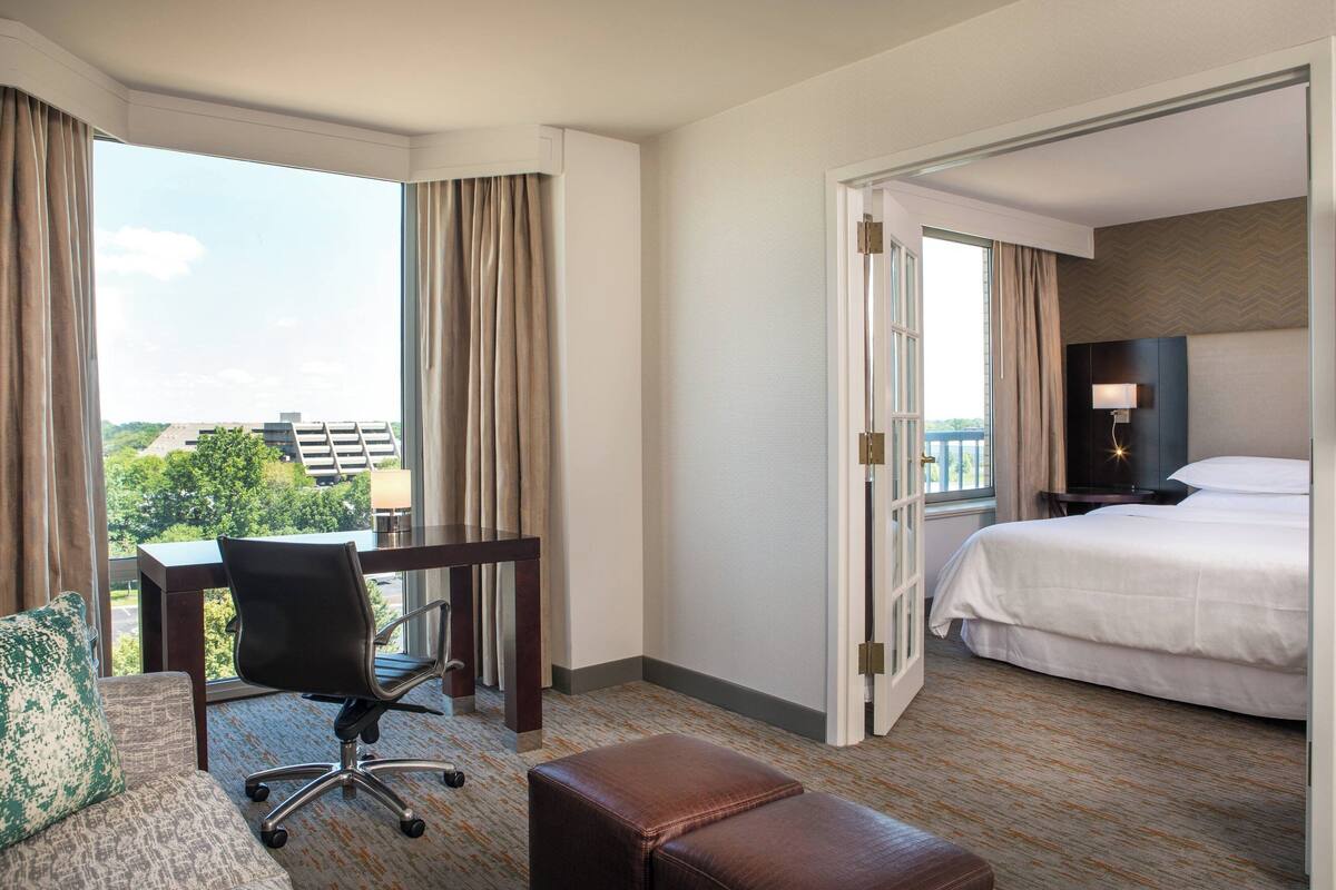 A hotel room with a bed, a desk, a chair, and a view of the city.