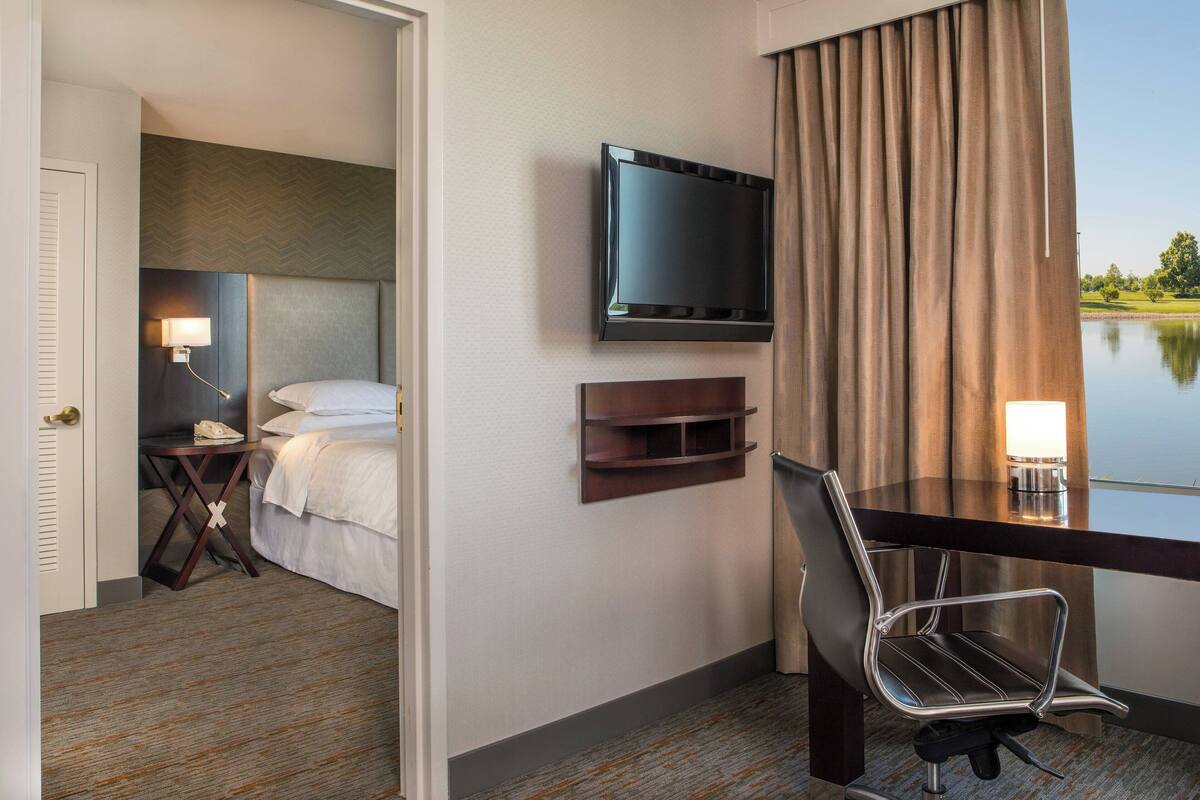 A hotel room with a bed, a desk with a chair, a television, a lamp, and a view of a lake.