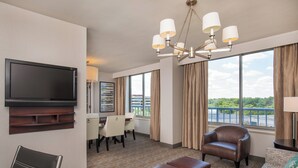 Habitación de hotel con televisor de pantalla plana, zona de comedor con mesa y sillas, sillón de cuero, sofá y ventana grande con vista a un puente y zonas verdes.