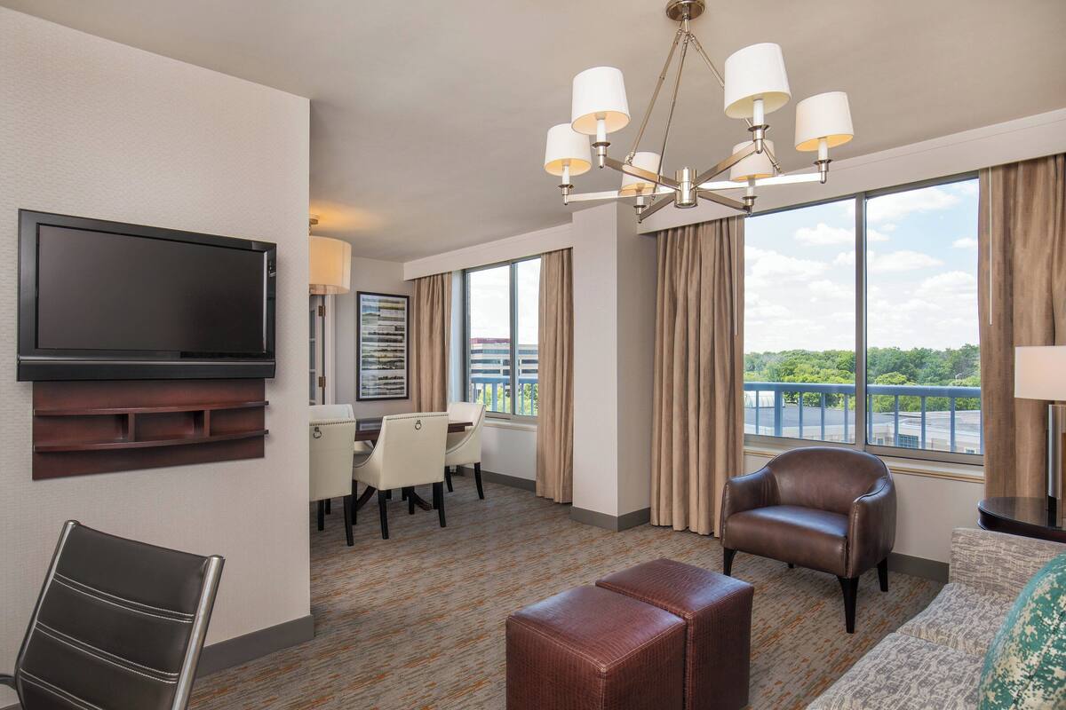 A hotel room with a flat-screen TV, a dining area with a table and chairs, a leather armchair, a sofa, and a large window with a view of a bridge and greenery.