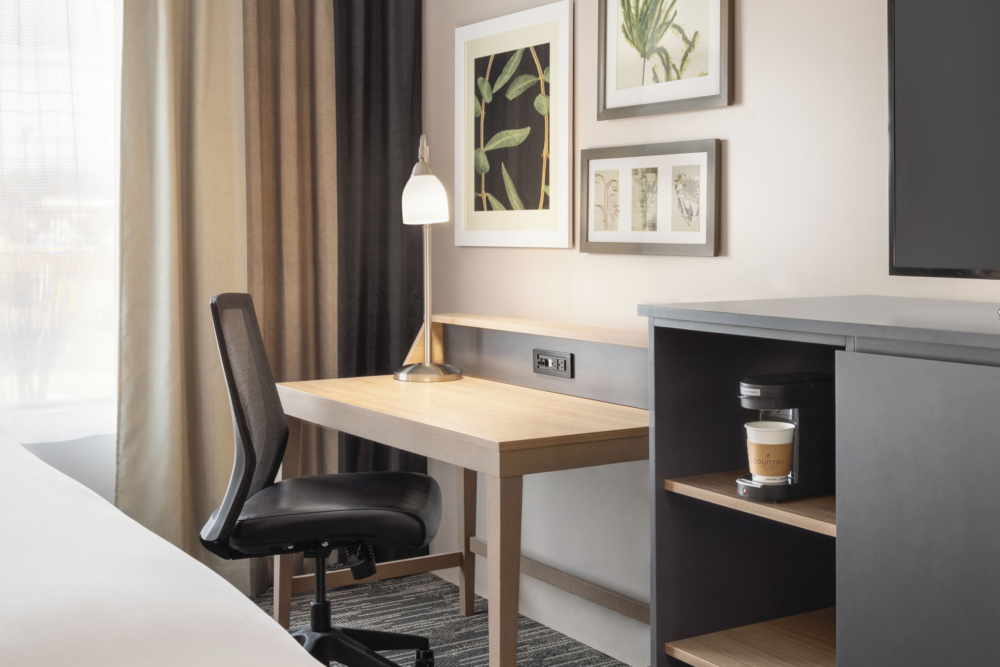 A hotel room with a desk, chair, coffee machine, and artwork on the wall.