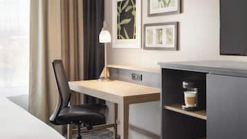 A hotel room with a desk, chair, coffee machine, and artwork on the wall.