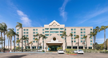A multi-story hotel with a central entrance, palm trees, and a clear blue sky.