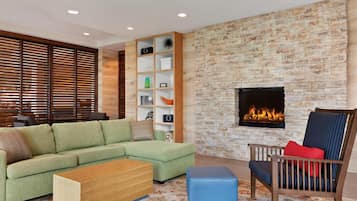 Un salon comprenant un canapé d’angle, une table basse en bois, un pouf bleu, une chaise en bois avec des coussins, une bibliothèque intégrée et un mur en pierre avec une cheminée.