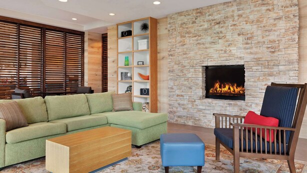 A living room with a green sofa, wooden coffee table, blue ottoman, and a chair with red cushions. A bookshelf is against the wall, and a fireplace is visible. - Country Inn & Suites by Radisson, San Diego North, CA (San Diego)