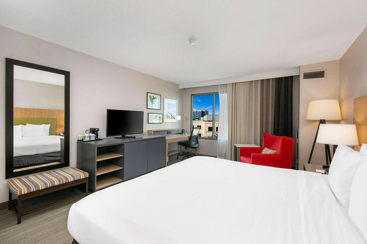 Une chambre d’hôtel avec un grand lit, un bureau avec un ordinateur, une télévision, un fauteuil rouge et un grand miroir.