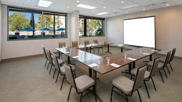 A conference room with a long table, chairs, a projector screen, and large windows with a view of outdoor umbrellas.