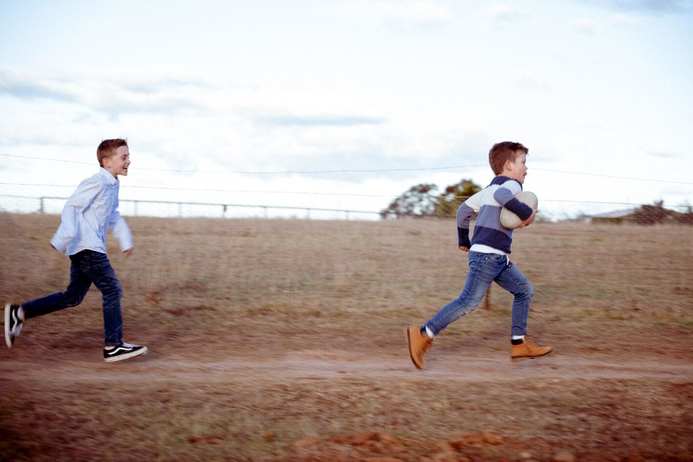 Two individuals running on a dirt path in an open field with a fence in the background.