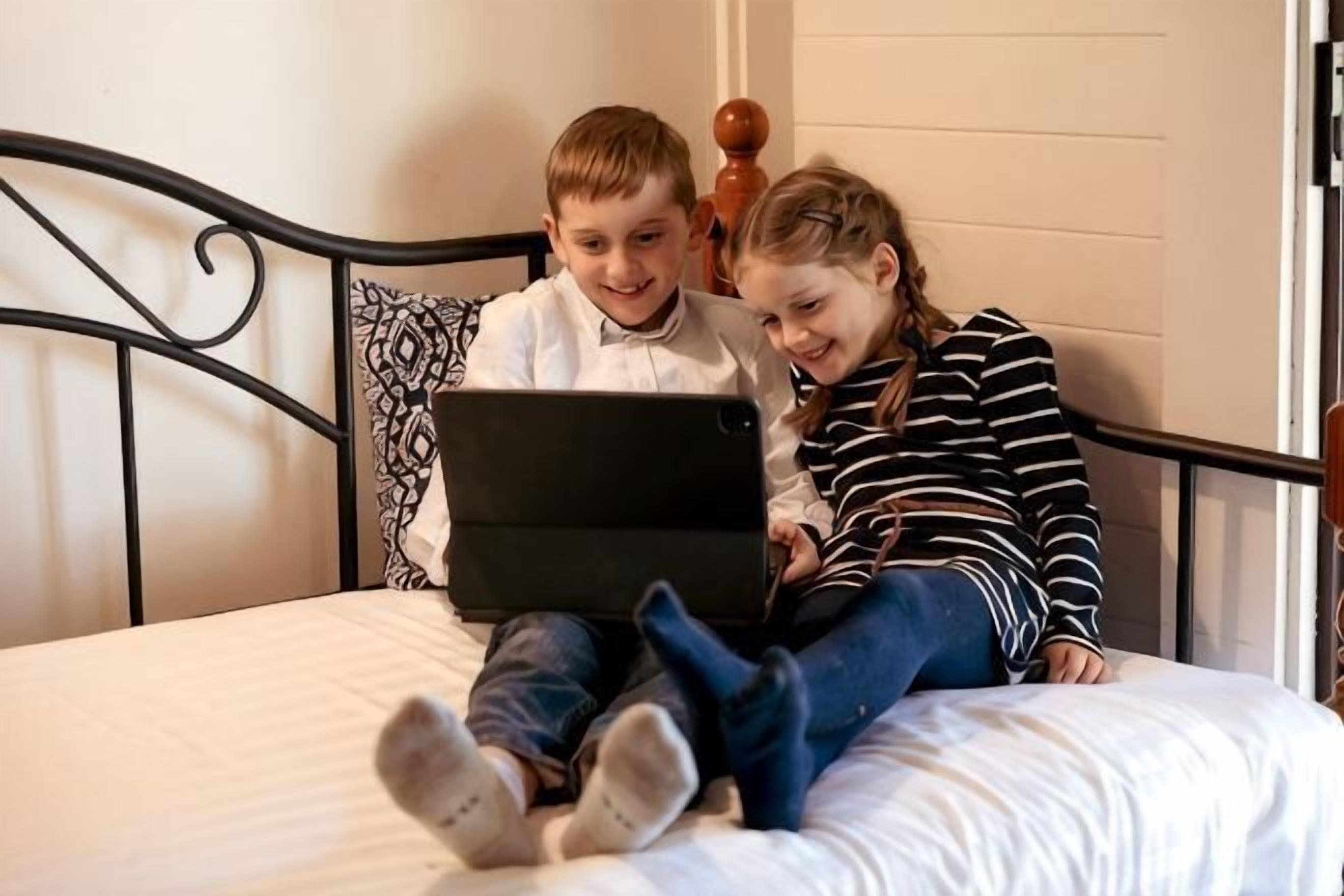 Two individuals on a bed with a laptop, a metal headboard, and a white wall.
