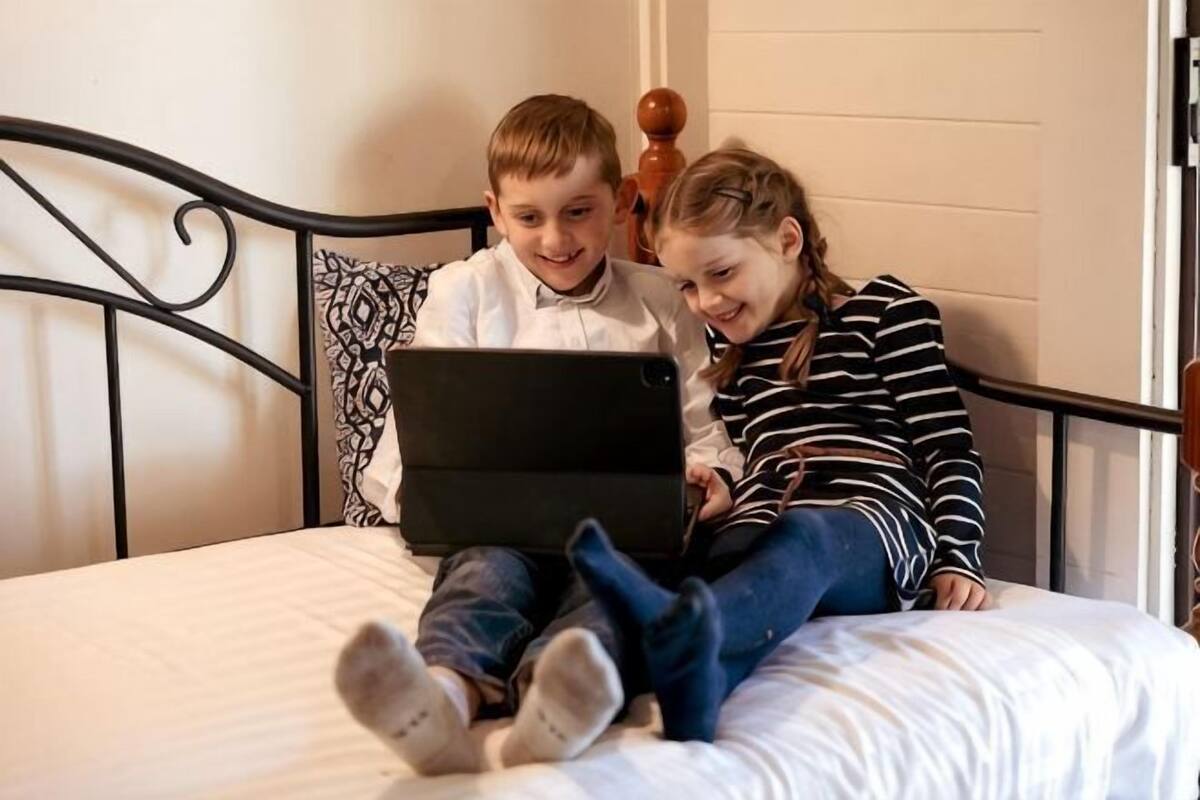 Two individuals on a bed with a laptop, a metal headboard, and a white wall.