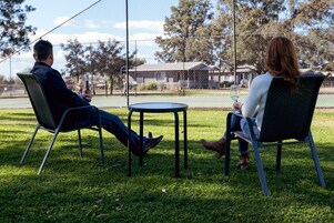 Dos personas sentadas en sillas frente a una pequeña mesa redonda en un espacio al aire libre con césped, una cerca y árboles al fondo.