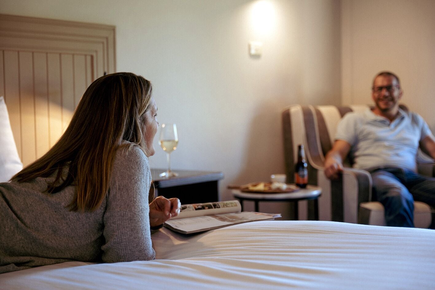 Une chambre d’hôtel avec un lit, une table de chevet sur laquelle se trouve un verre de vin, une assiette de nourriture et une bouteille de bière.