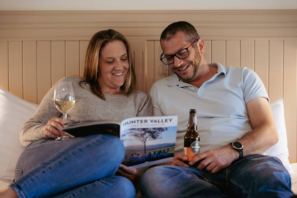 A couple sitting on a bed with a book titled "Hunter Valley" and a bottle of wine.