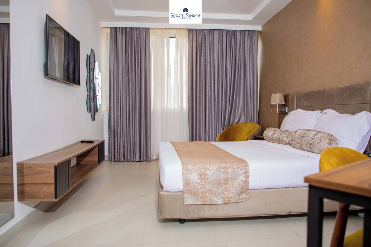 Een moderne hotelkamer met een bed, een houten console en een televisie die aan de muur is bevestigd, plus een groot raam met gordijnen.