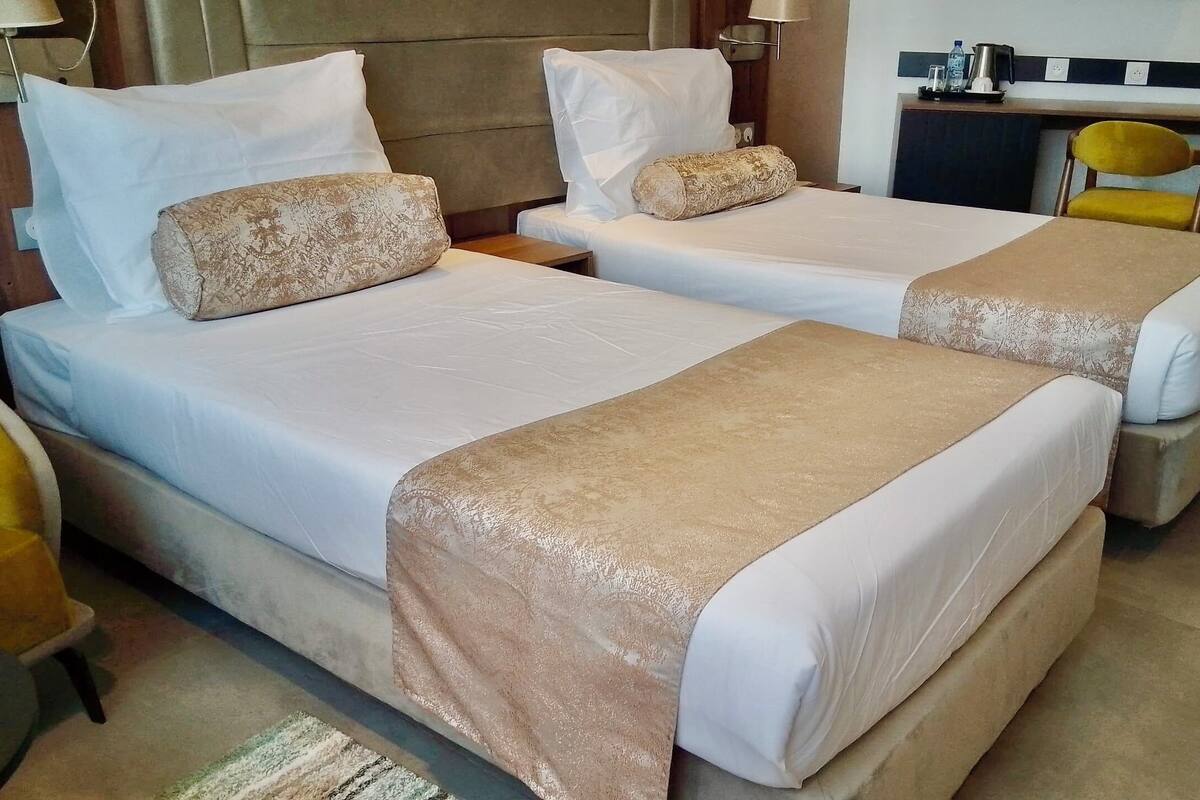 Een hotelkamer met twee bedden, elk voorzien van een beige hoofdbord en witte beddengoed. In de hoek staat een klein tafeltje met een lamp en een stoel.