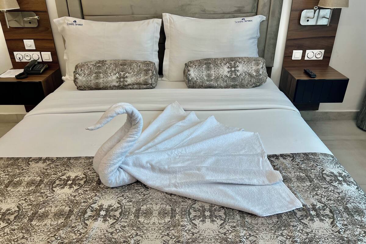 Een opgemaakt bed met een handdoek in de vorm van een zwaan, twee gestreepte kussens, bedlampjes en een nachtkastje met een telefoon en afstandsbediening.