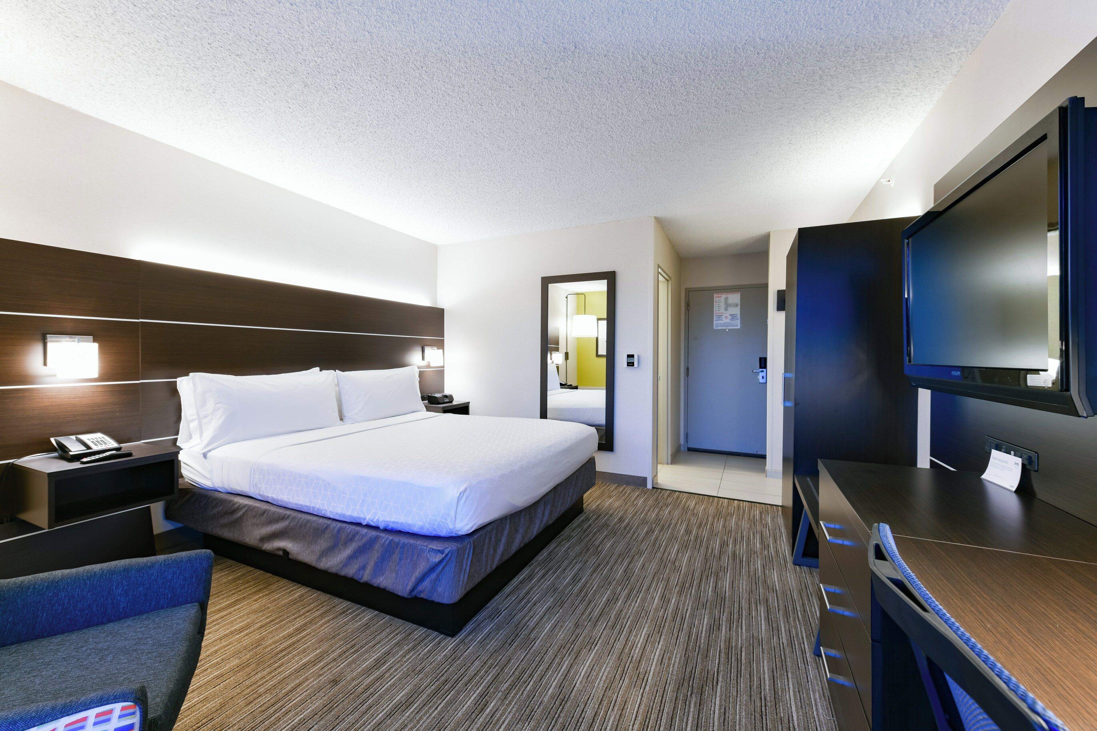 A hotel room with a large bed, bedside tables, a desk with a chair, a flat-screen TV, and a door leading to another room.