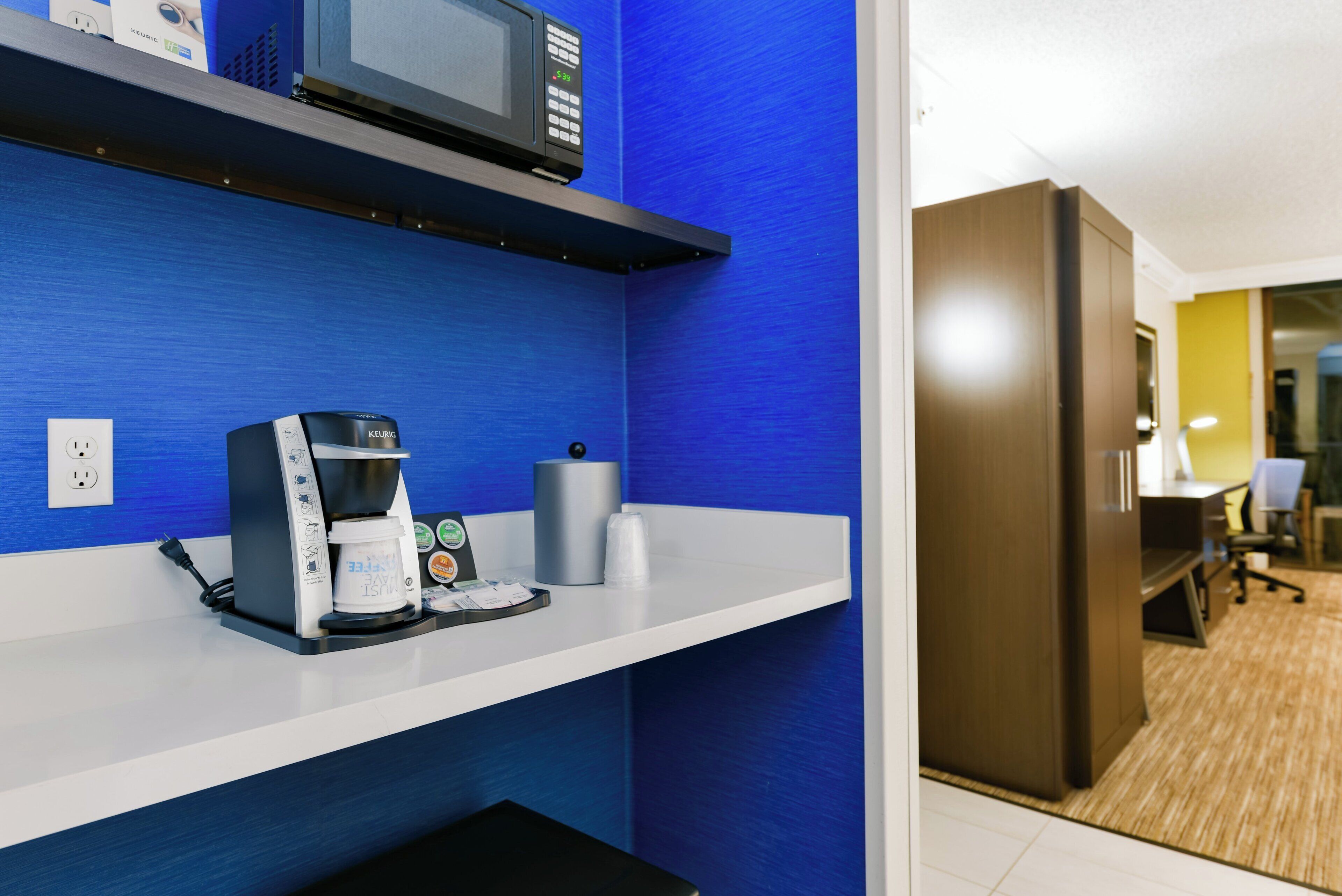 Una kitchenette con cafetera, microondas y hervidor sobre una pared azul.