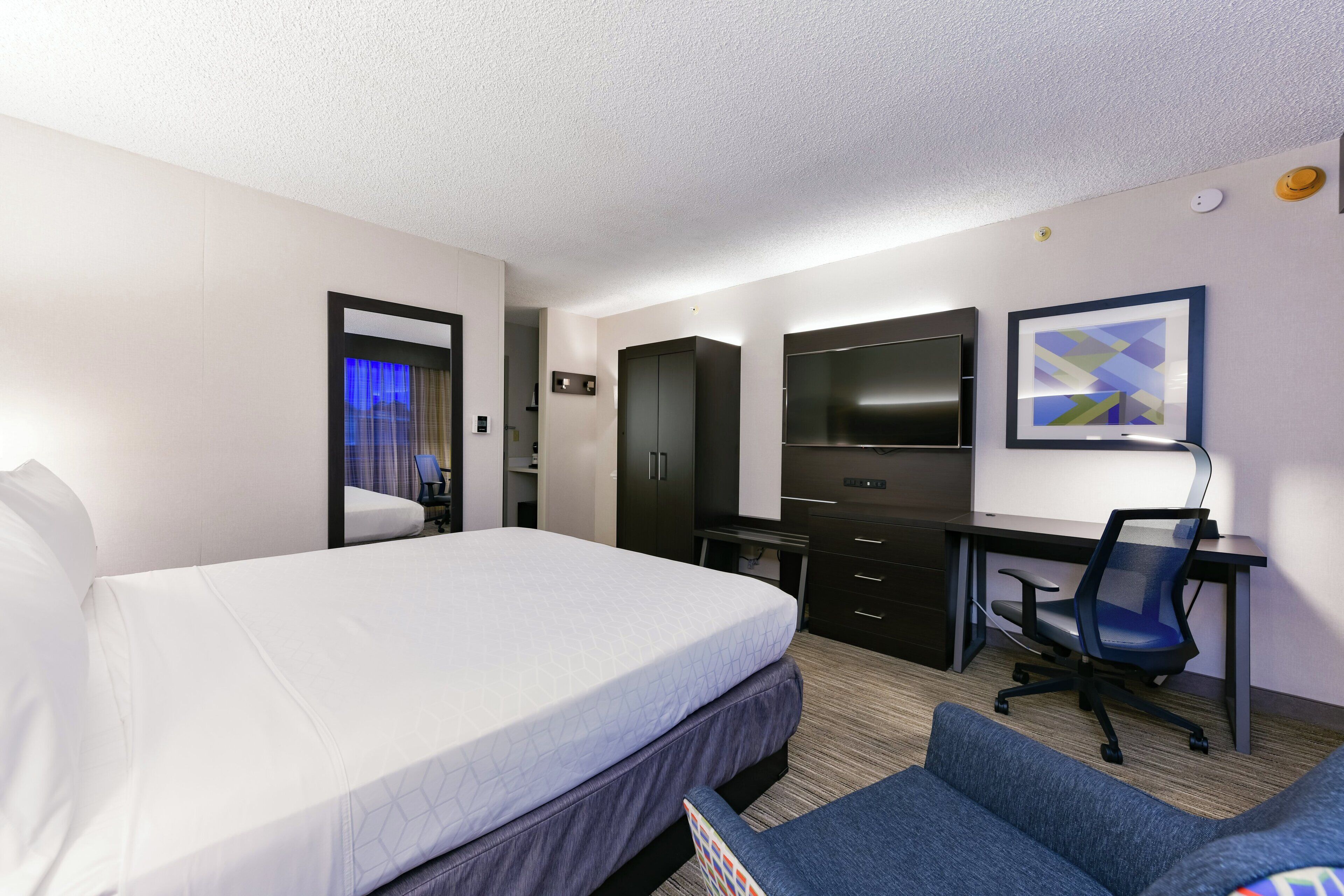 Ein Hotelzimmer mit einem Bett, einem Schreibtisch mit Stuhl, einem Fernseher und einem gerahmten Bild an der Wand.