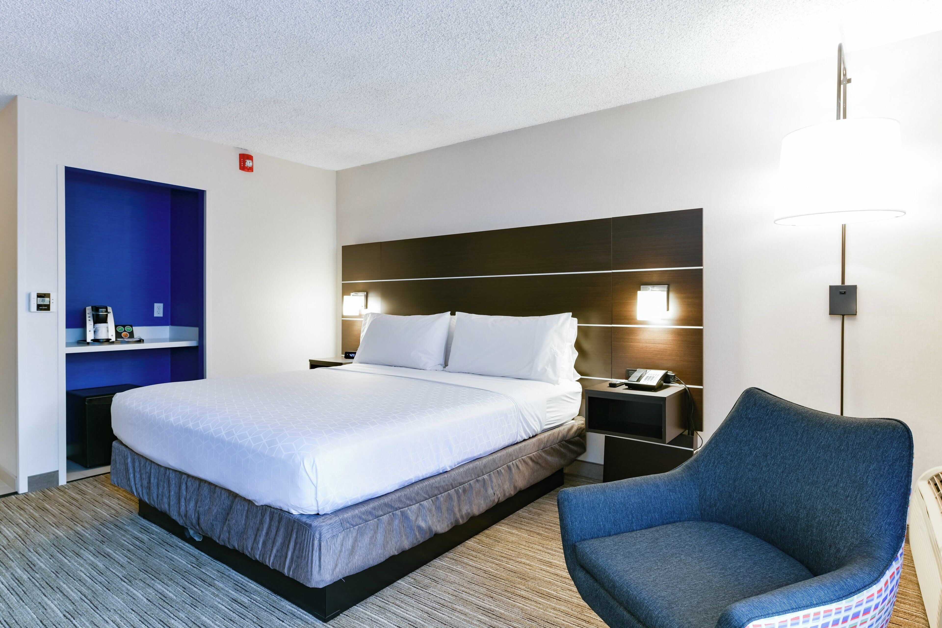 A hotel room with a bed, a chair, a nightstand, and a wall-mounted lamp.