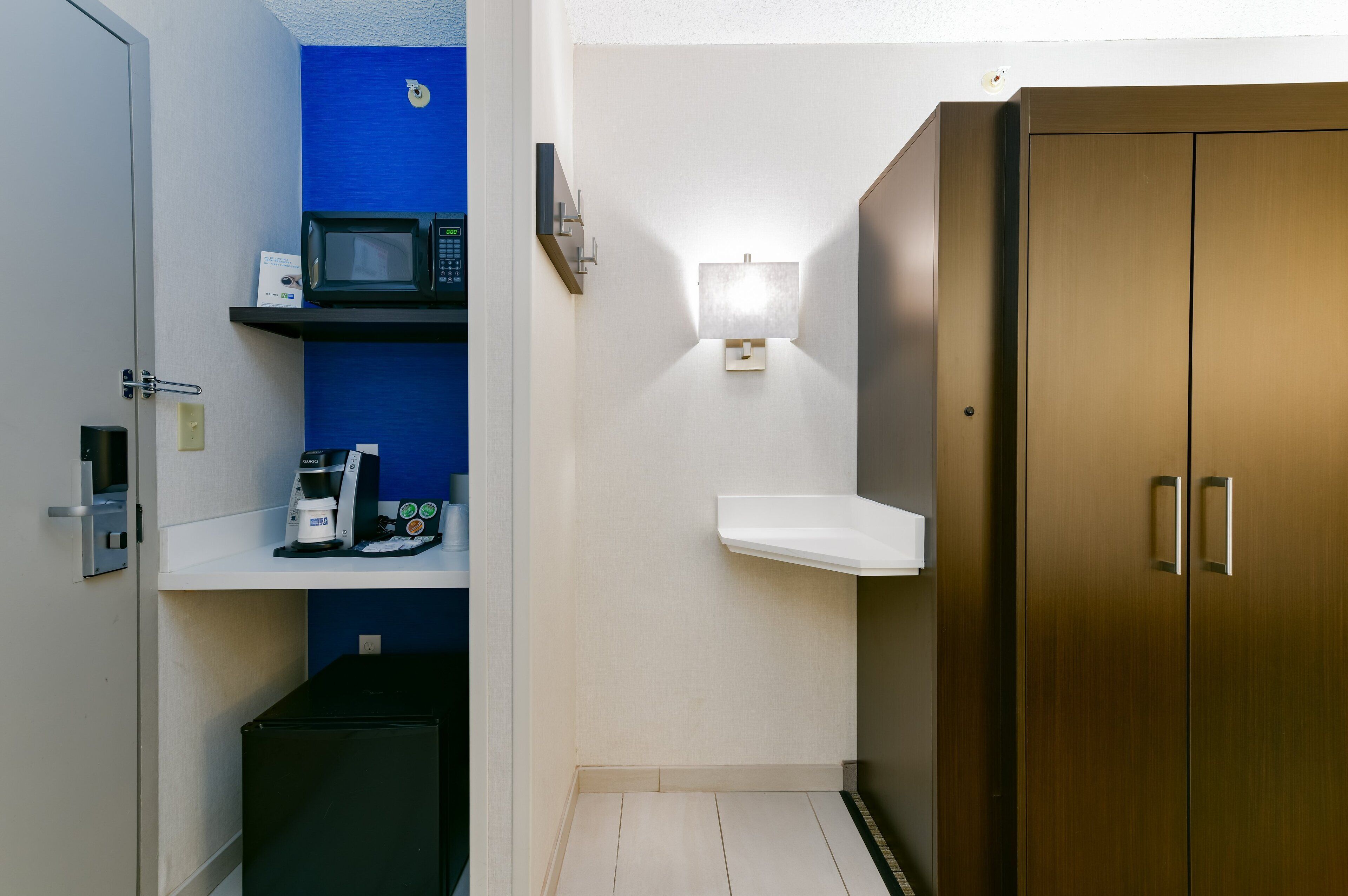 A compact hotel room with a microwave, coffee maker, and a small shelf.