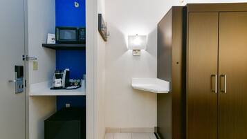 A compact hotel room with a microwave, coffee maker, and a small shelf.