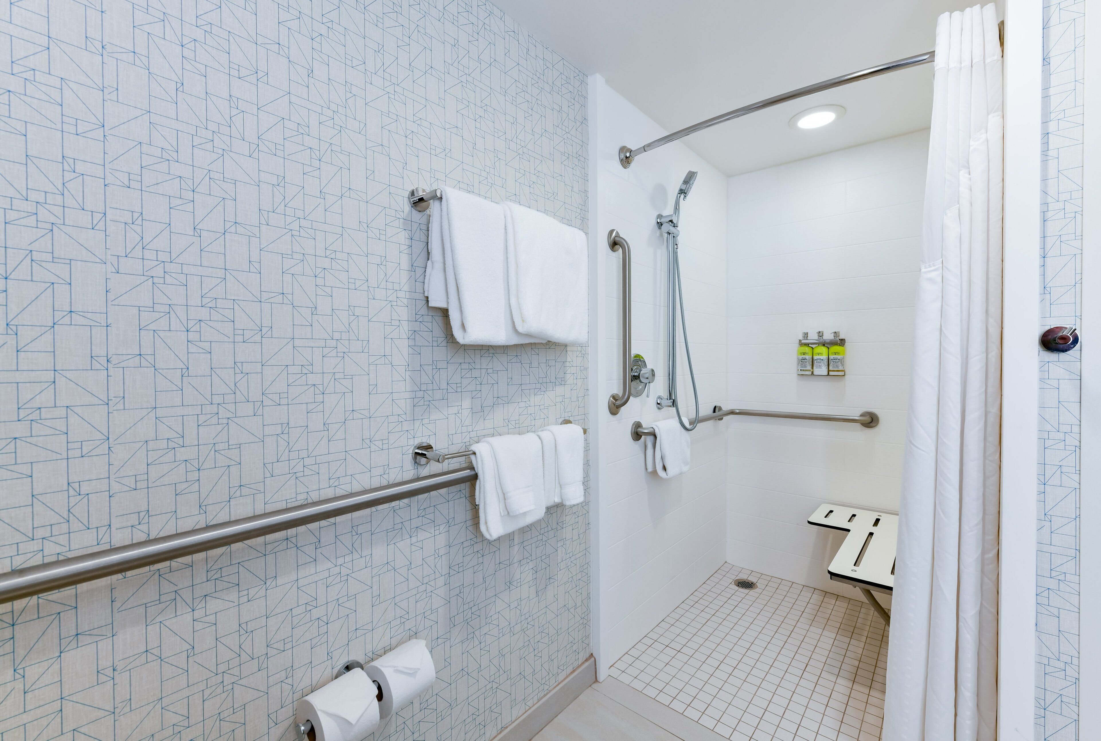 Un baño moderno con un patrón geométrico de azulejos, ducha a ras de suelo, barras de apoyo y un inodoro con barra de seguridad.