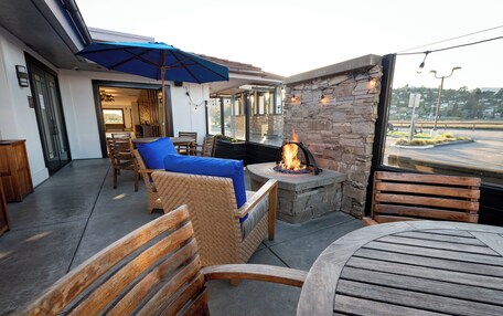 An outdoor patio with a stone wall, a fire pit, wooden furniture, and a blue umbrella.