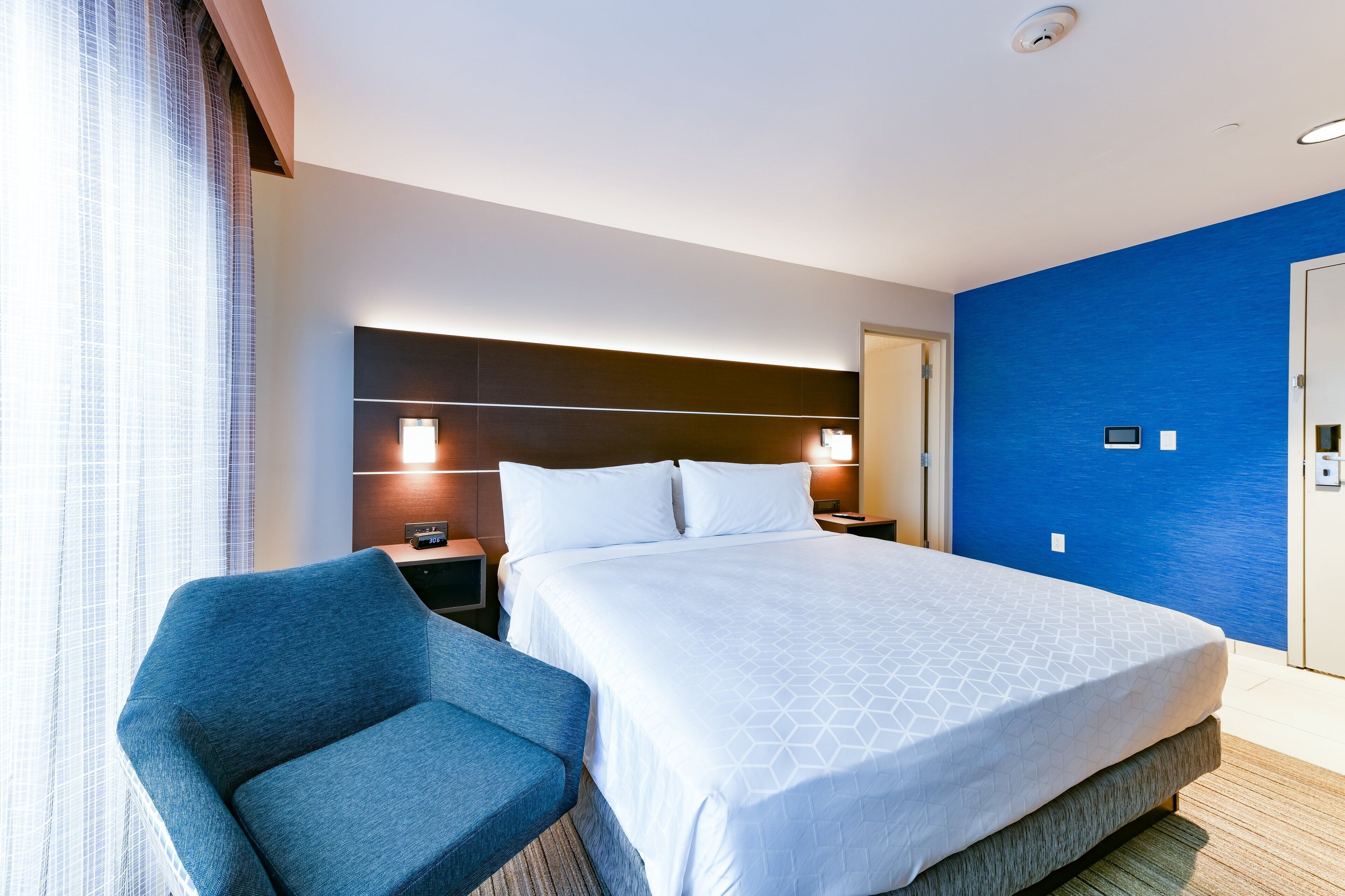 Een hotelkamer met een groot bed, een blauwe fauteuil, een nachtkastje en een aan de muur bevestigde televisie.