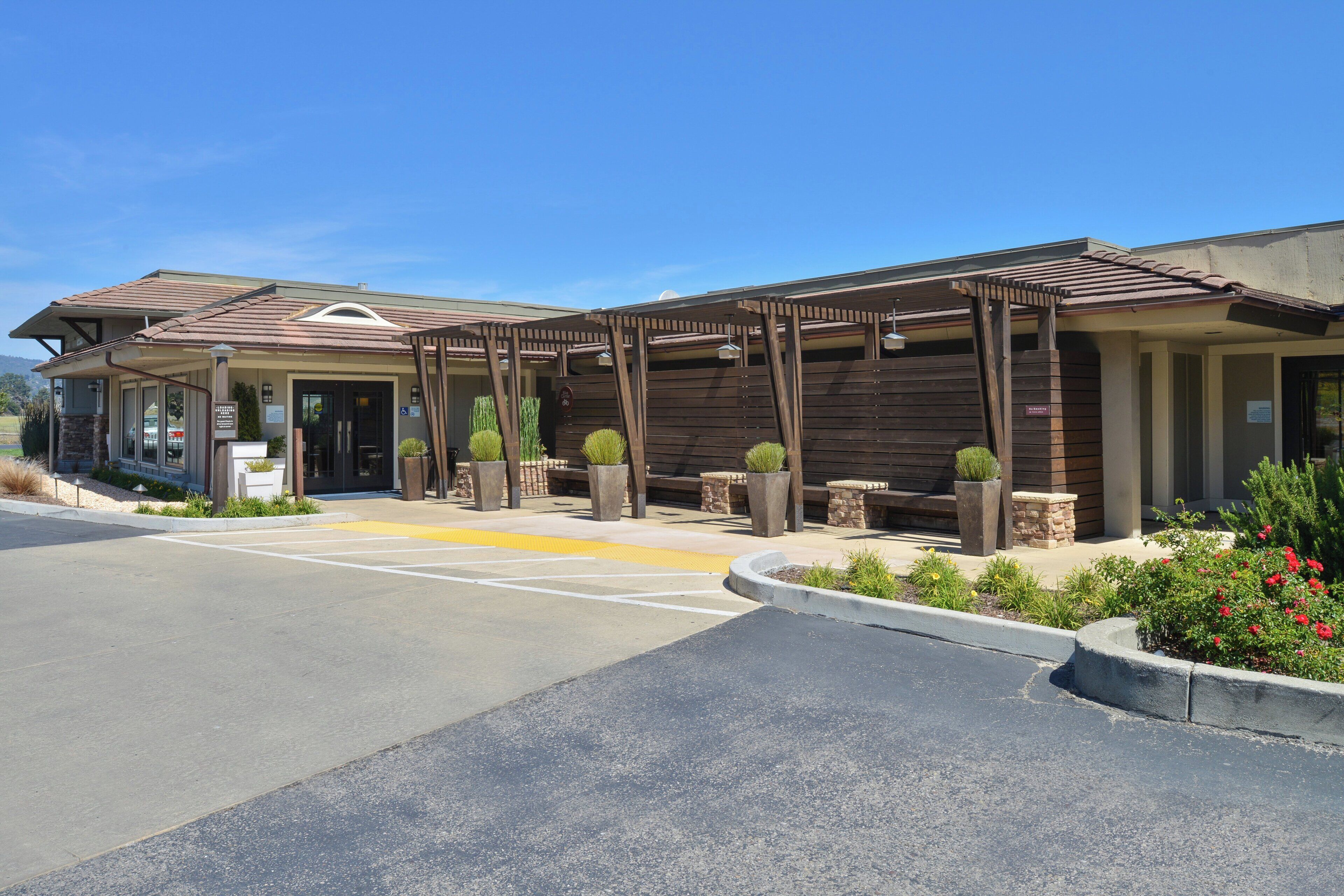 A hotel entrance with a covered outdoor area, a parking lot, and landscaped gardens.
