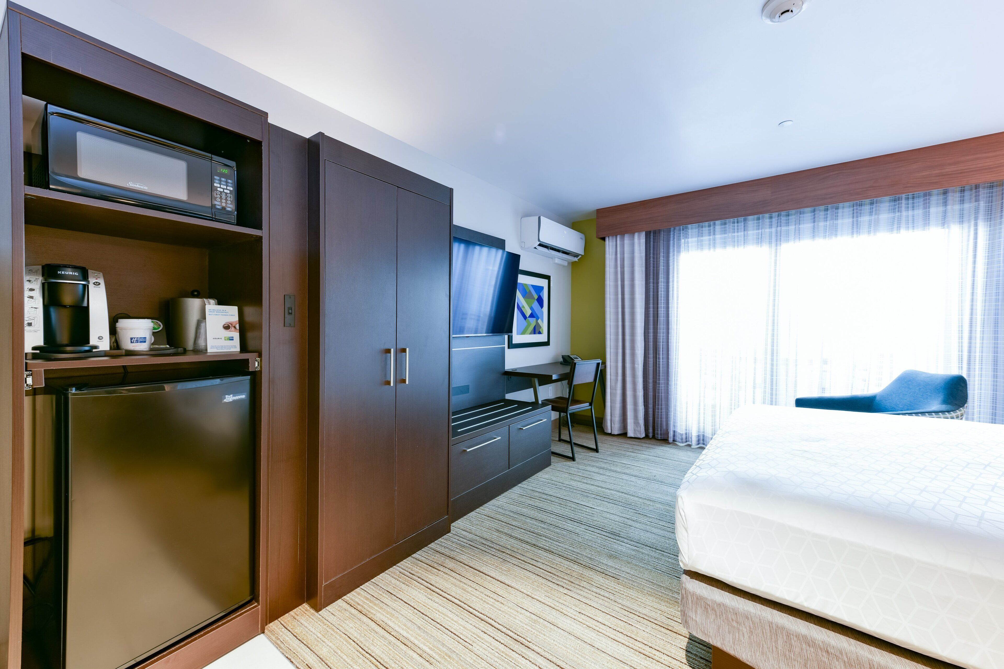 A hotel room with a bed, a desk with a chair, a microwave, a coffee machine, and a mini-fridge.