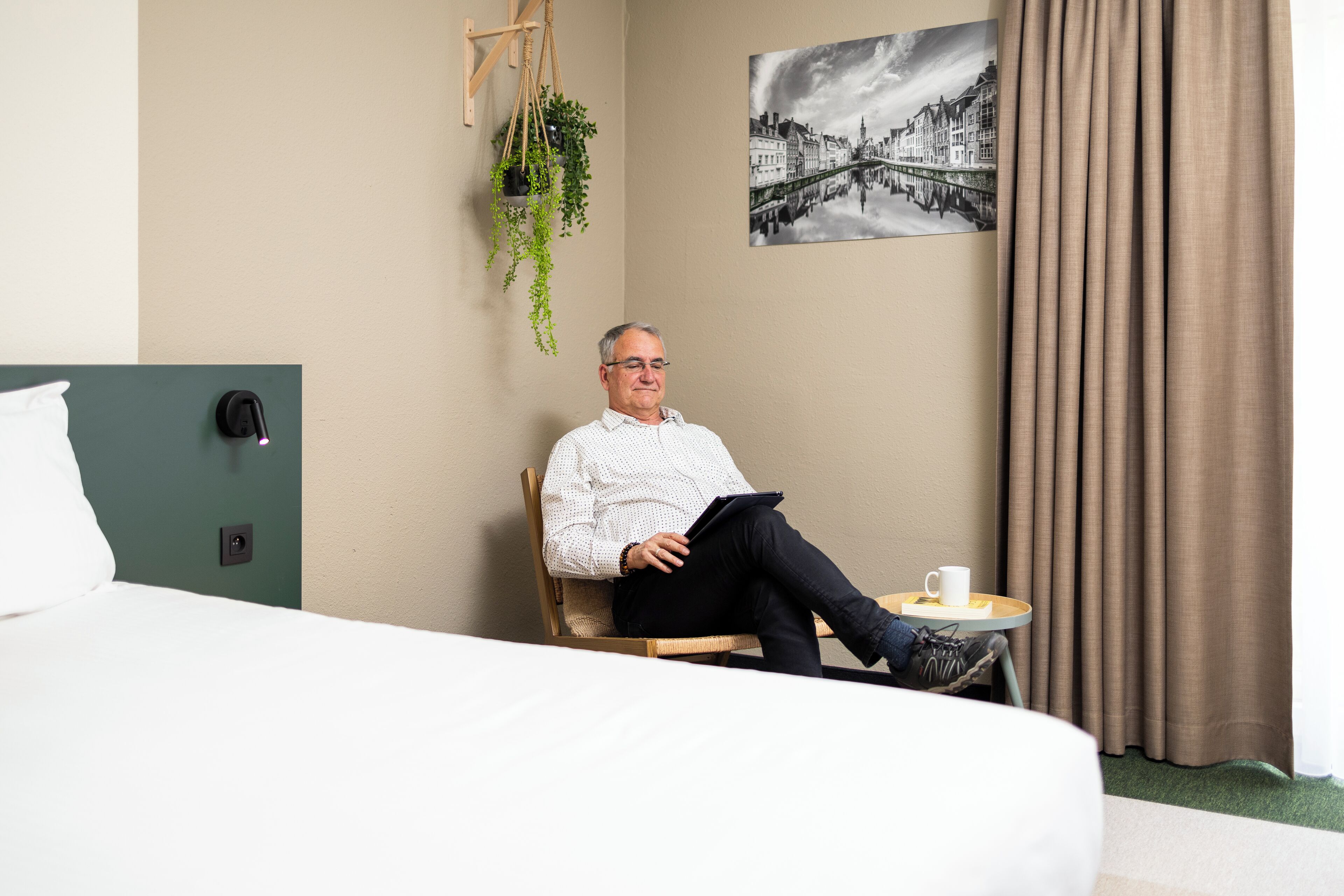 A man sitting on a chair in a hotel room, holding a tablet. There is a bed with a white pillow and a green headboard. A small table with a cup and a plant is visible.