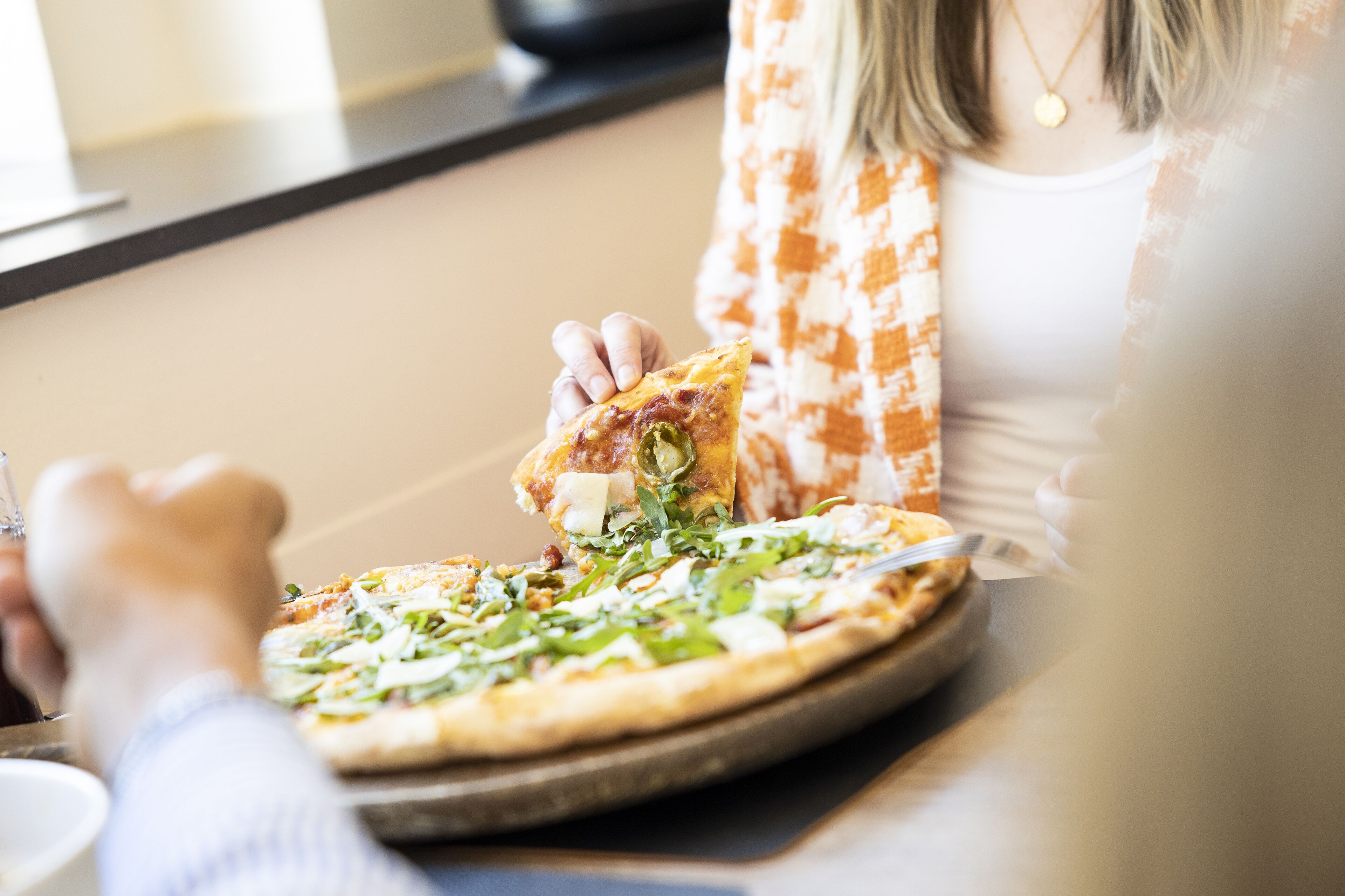 A person holding a slice of pizza with toppings, sitting at a table with a pizza in front of them.