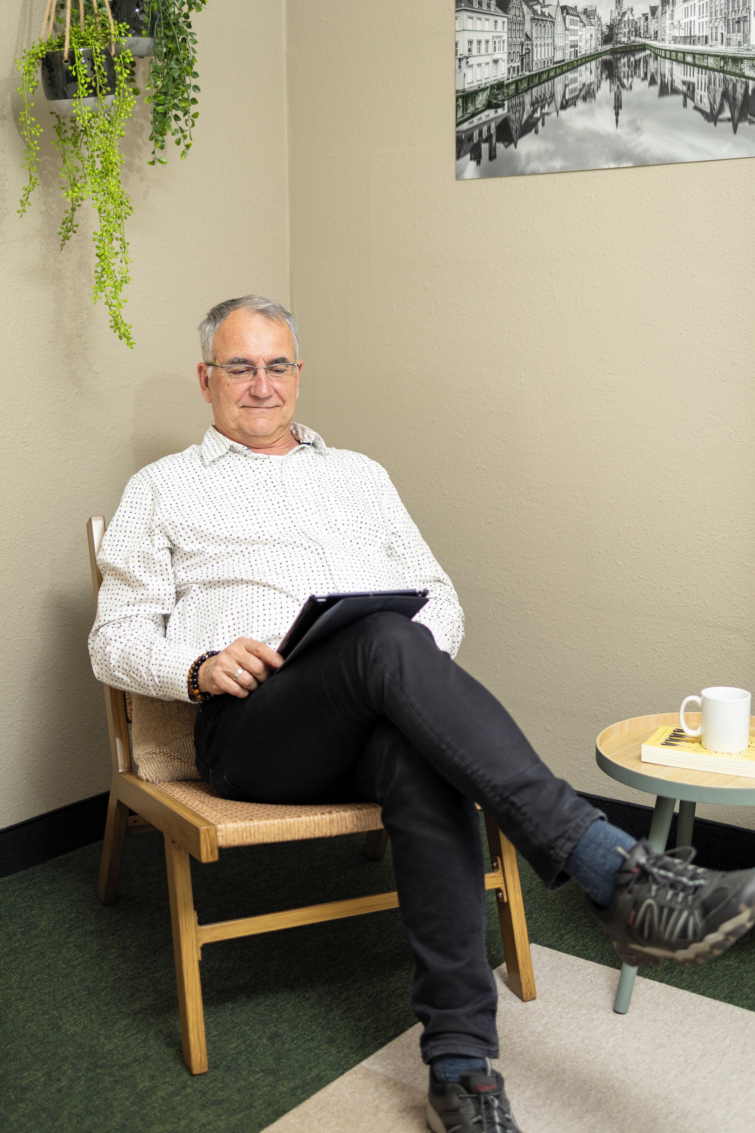 A man sitting on a chair in a room with a green carpet, holding a clipboard. There is a small round table with a white mug on it.