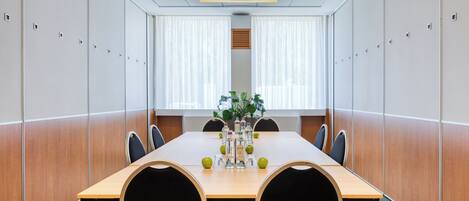 Un salón de conferencias con una larga mesa, sillas, una planta y un florero con manzanas y botellas.