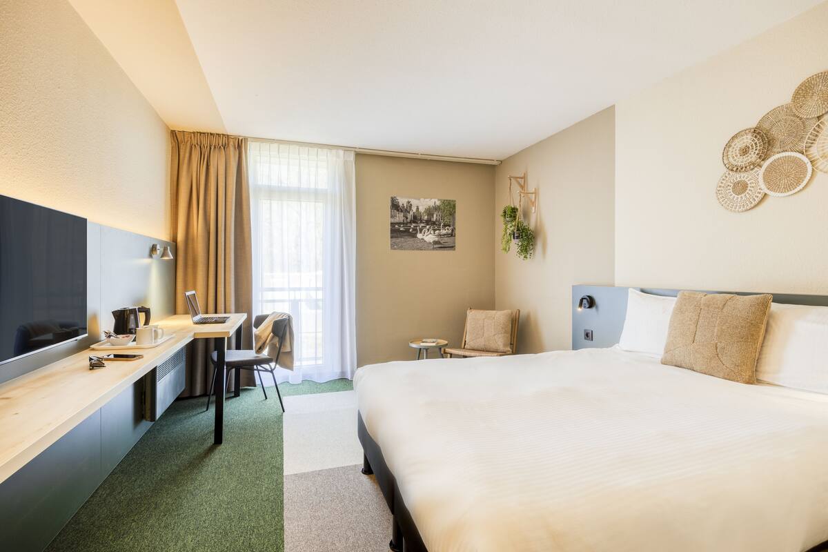 Habitació d'hotel amb un llit gran, un escriptori amb un ordinador portàtil, una cadira, un televisor i una finestra amb cortines.