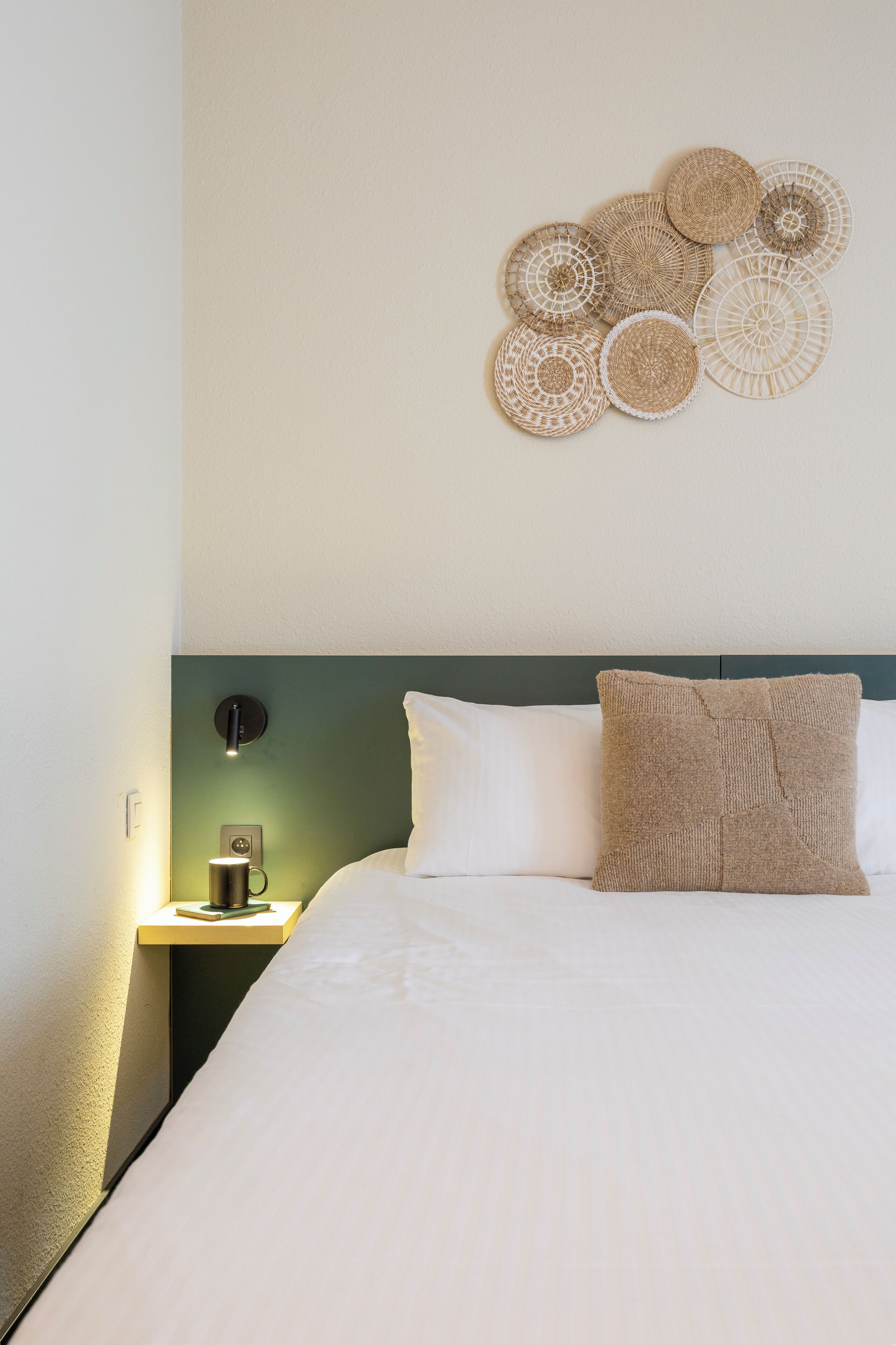 A neatly made bed with a white comforter and a textured throw pillow, a bedside table with a lamp, and a wall-mounted decorative item.