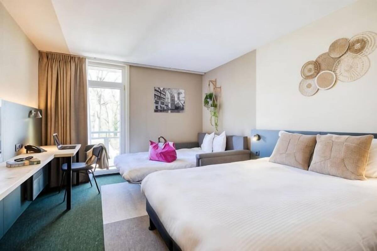 Habitació d'hotel amb un llit, escriptori, cadira, finestra i paret amb un objecte decoratiu.