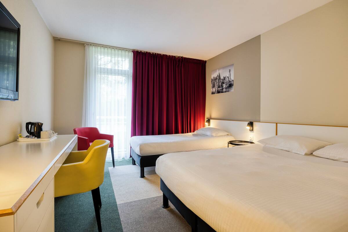 Une chambre d’hôtel avec deux lits, un bureau, une chaise, une fenêtre avec des rideaux rouges et un tableau accroché au mur.