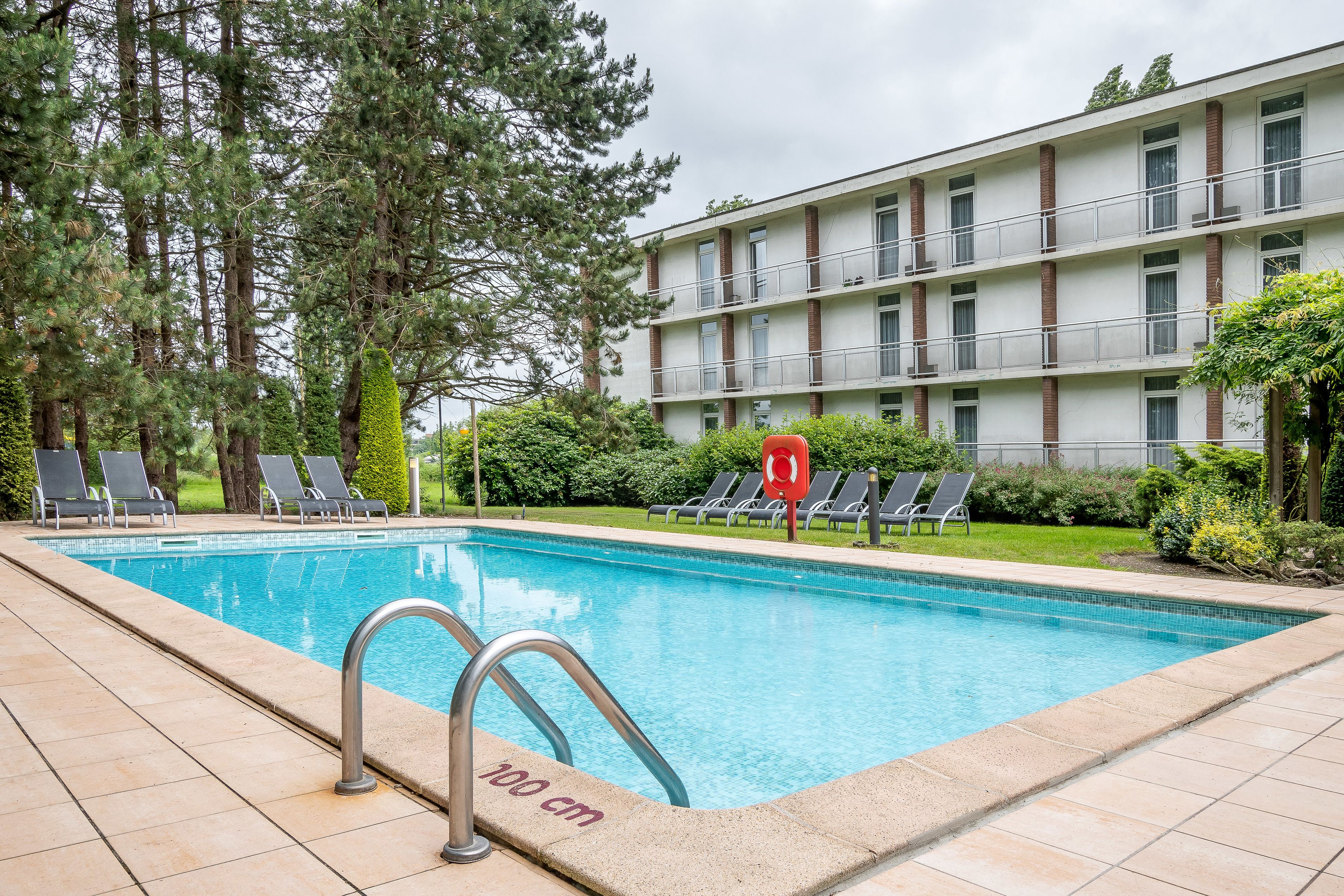 A swimming pool with a 100 cm depth marker, surrounded by lounge chairs and a multi-story building with balconies.