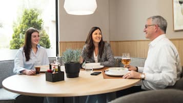 Three people seated at a round table with a potted plant, a glass of beer, and plates of food.