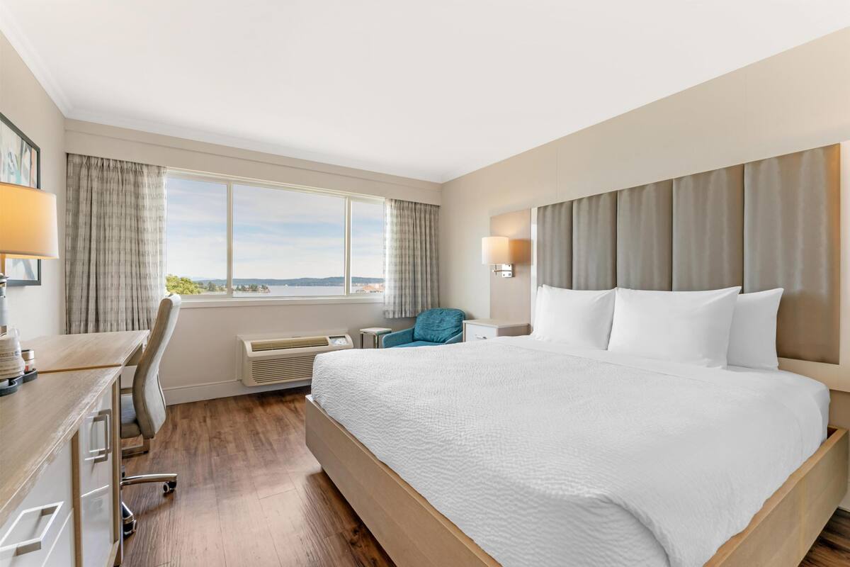 Ein Hotelzimmer mit einem großen Bett, einem Schreibtisch, einem Sessel, einem Fenster mit Vorhängen und Blick ins Freie.