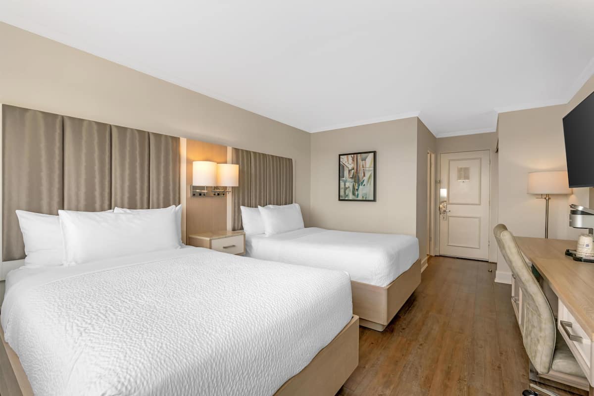 A hotel room with two beds, a desk, a chair, a TV, and a door.
