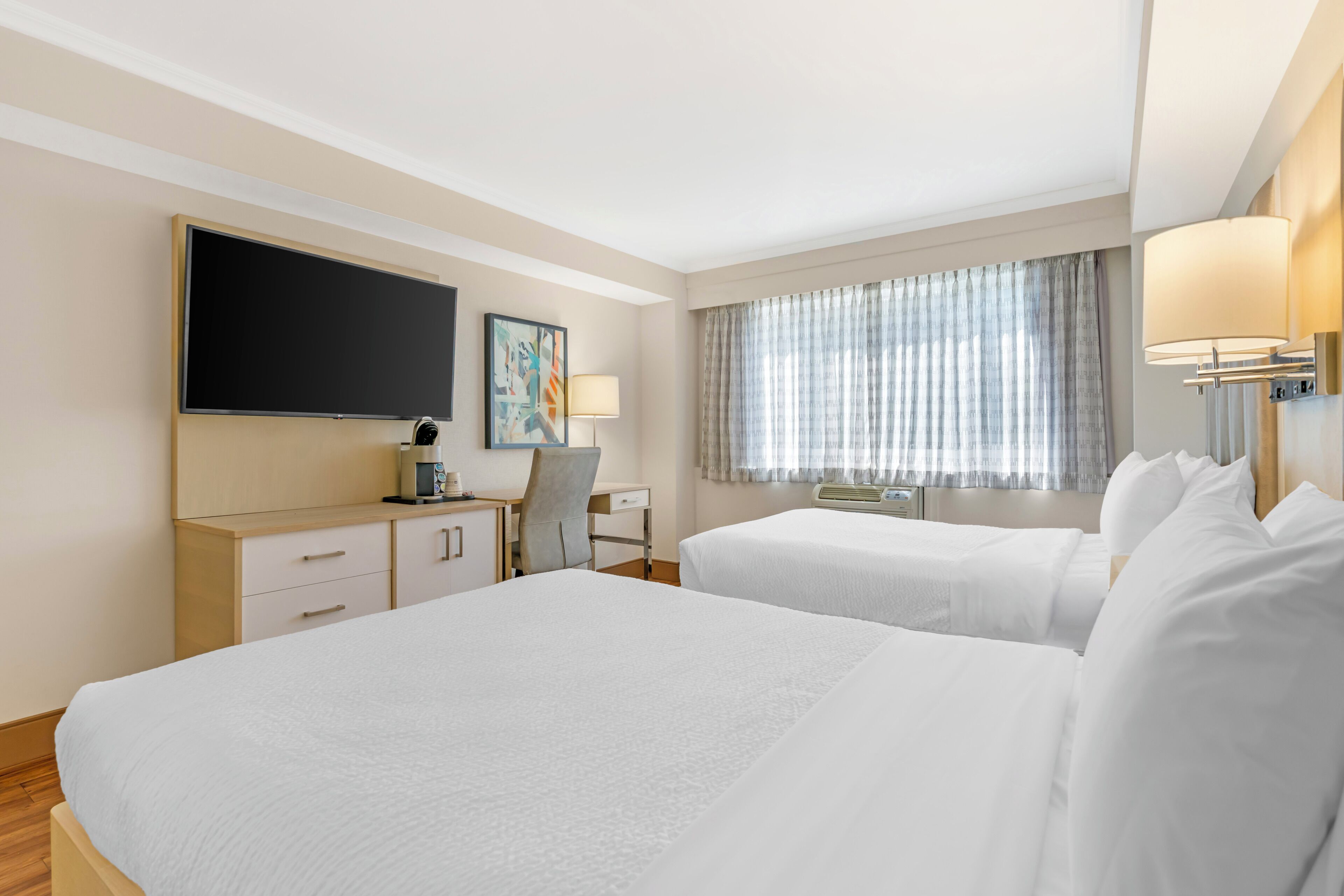 A hotel room with two beds, a desk, a chair, a flat-screen TV, a painting, a window with curtains, and a lamp.