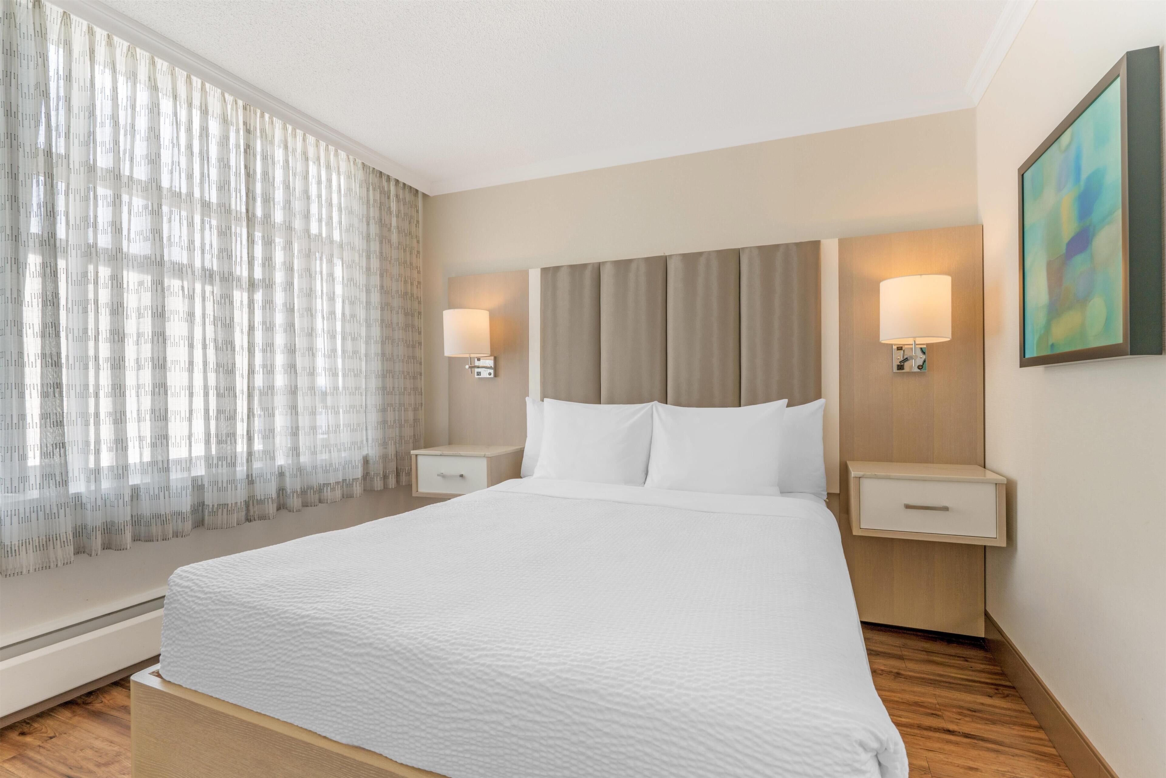 A hotel room with a large bed, two bedside tables with lamps, a window with sheer curtains, and a wall-mounted abstract painting.