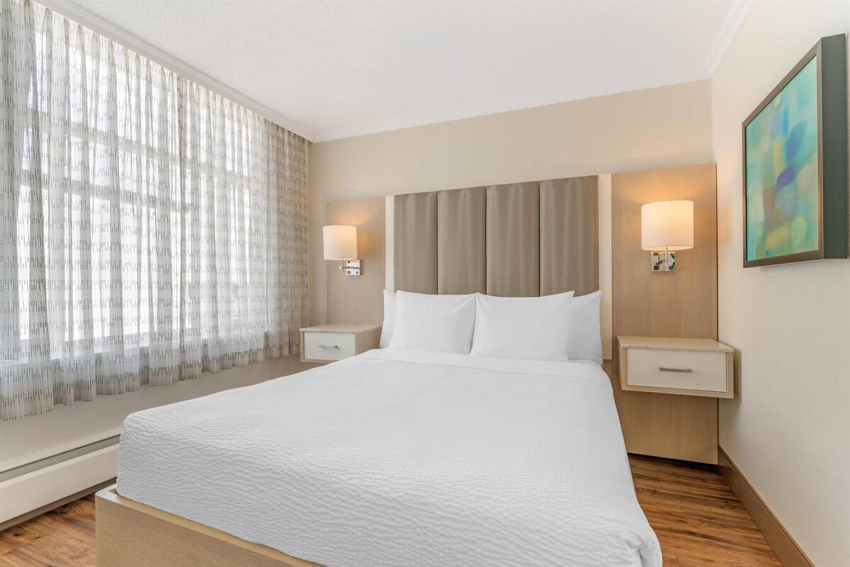 Ein Hotelzimmer mit einem großen Bett, zwei Nachttischen mit Lampen, einem Fenster mit durchsichtigen Vorhängen und einem an der Wand hängenden abstrakten Gemälde.
